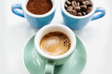 Three cups of coffee: ground coffee (powder), coffee beans and freshly made espresso in colorful ceramic cups (mint green and light blue) on the white background
