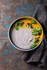 Mushroom cream soup in black ceramic bowl served with turmeric powder, forest mushrooms, greens on black textile napkin over old dark wooden background. Top view