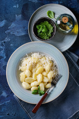 Blue plate of hot creamy potato gnocchi served with grated parmesan, fresh basil, pesto sauce, olive oil, fork on textile napkin over dark blue concrete background. Flat lay with space