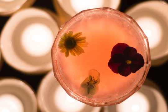 Pink Cocktail Garnished With Flowers From Above. Candlelit Alcoholic Drink In Margarita Glass With Pansies And Daisy Floating