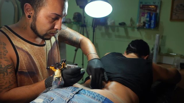 Portrait of man tattoo master with dreadlocks showing process of creation tattoo on female body under the lamp light. Slow motion. Professional artist working in sterile gloves