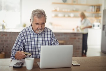 Fototapeta premium Senior man paying bills online on laptop in kitchen