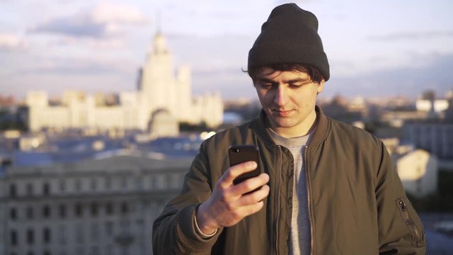 Young man wearing a khaki jacket and a beanie is texting on his smartphone while standing on a roof of a building. Locked down real time medium shot