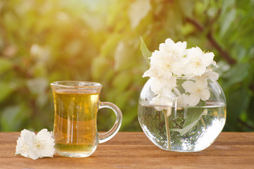 Transparent mug of tea and a vase with jasmine on a wooden table, greens on the background