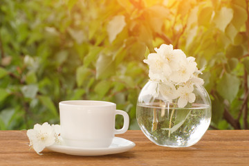 White mug of tea and a vase with jasmine on a wooden table, greens on the background