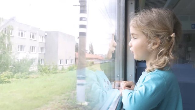 Train departs from station. Little curious girl looking out of window in train. Drops of rain on glass. It's raining outside, child reflecting in glass. Sad child looks out the window.