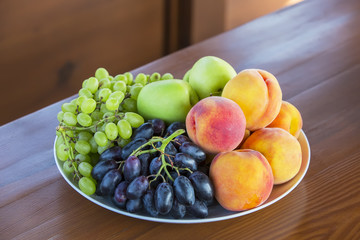 Fresh fruits on a white plate on wooden background. Ripe peaches, grapes branch, green apples.
