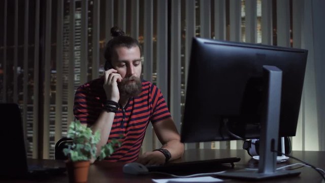 Bearded man sitting at computer and talking on phone in the office at night.