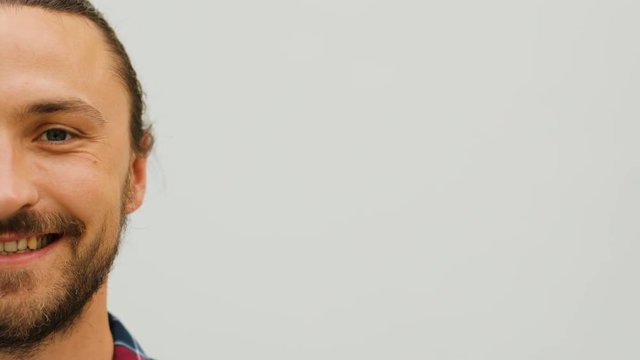 Close up shot of left half face of attractive serious man with beard on the white wall background.