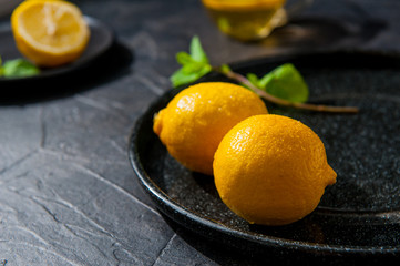 Fresh ripe lemons with water drops and mint leaf on the black plate and cup with herb tea on the dark stone background. Selective focus. Space for text