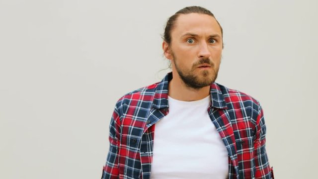 Close up shot of causasian young man in casual shirt making funny faces on the camera on white wall background.