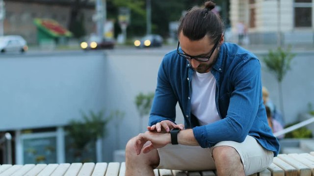 Portrait of caucasian young man using smart watch on his hand while relaxing on bench on the city street background.