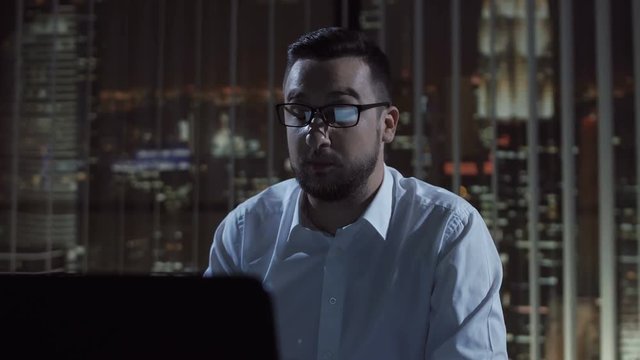 Thoughtful man in glasses sitting at laptop and working late in office making videocall.