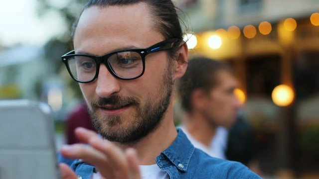 Close up portrait of young man with the glasses in the denim shirt using tablet for chatting with friends in the city street. Side view.