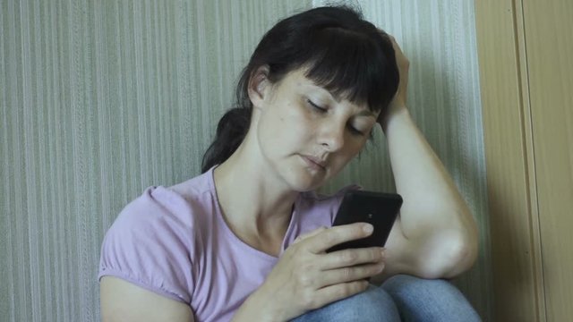 Sad young woman sits on the floor with a smartphone in her hand. A girl in a depression uses a smartphone, writes a text message, sitting on floor in corner of  room.