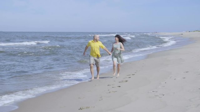 Happy lovely couple running along the seacoast barefoot and enjoys herself. Beach with white sand. Outgoing coastal line. Slow Motion.