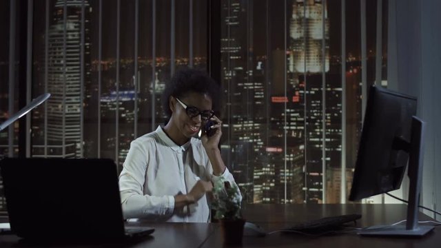 Cheerful black woman smiling and talking phone at computer in office in the evening.