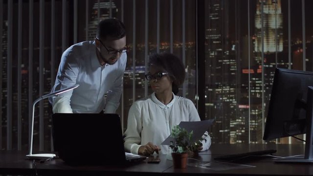 Black and white woman and man at desk in office doing paperwork in the evening.