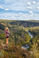 Naklejka premium Young blonde woman tourist on a cliff taking pictures of the autumn landscape with the river Berd on old film camera
