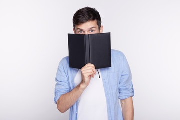 Studio shot of European blue eyed businessman or student dressed casual, hiding face behind notebook while standing isolated on white background,looking humorously at camera. People, business, career.