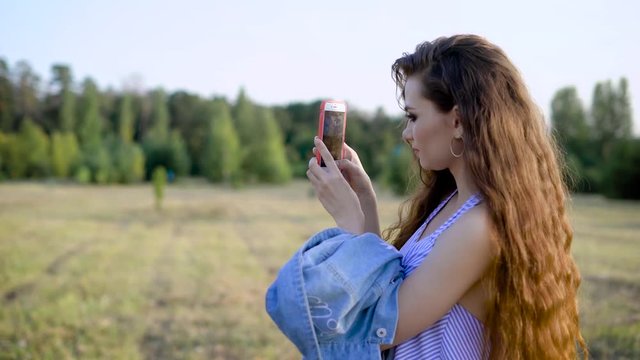 An adult girl with smoky eyes and curly hair takes photos of nature in order to put on social networks through the Internet, a lady walks through the park