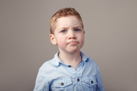 Closeup portrait of cute adorable blond redhead Caucasian little boy in blue shirt standing in studio on light white background. Child kid looking in camera with funny sceptical face expression