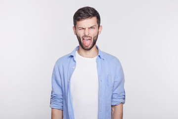 Fototapeta premium Displeased and bearded young handsome blue eyed man employee wearing white shirt grimacing, making mouths, sticking out his tongue at camera trying to tease someone, acting like little naughty child.