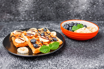 On a dark background, a plate with homemade Belgian waffles, poured with chocolate sauce, with sliced banana, blueberries and mint.