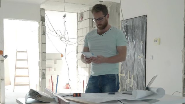 Male architect with cellphone and tablet checking wall with level at new apartment 
