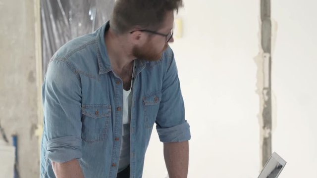 Male architect with blueprints checking wall with level at new apartment 
