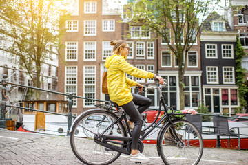 Fototapeta premium Młoda kobieta w żółtym płaszczu przeciwdeszczowym z torbą i kwiatami, jazda na rowerze w mieście Amsterdam