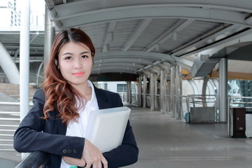 Portrait of confident young Asian business woman standing and holding document folder in her hands at walkway against copy space background.