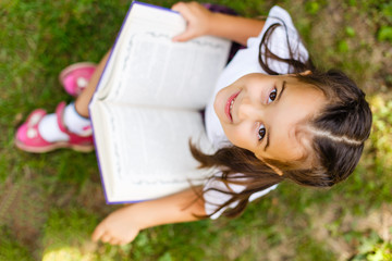 Portrait of little cute girl reading book, sitting at summer green park.