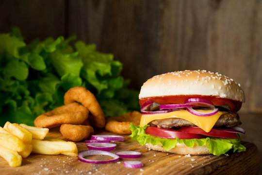 Cheeseburger, french fries and onion rings on wooden cutting board over wooden background. Closeup view, selective focus. Fast food concept