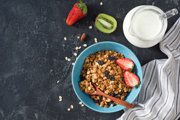 Homemade granola with raisins and nuts in blue bowl decorated with strawberries and jug of milk on dark stone background. Table top view and copy space for text, horizontal composition