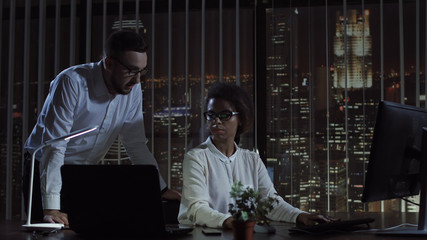 Black and white woman and man working together at computer in office at night.