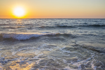 Panorama del sole che tramonta a mare. L'acqua è mossa e le onde a contatto con la sabbia formano la schiuma. Il mare è agitato e il cielo è arancione per via del crepuscolo.