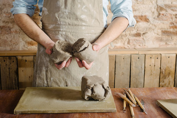 potter, workshop, ceramics art concept - standing young brunette man dressed in an apron, male hands knead the fireclay, a ceramist with raw materials on wooden table with sculpting tools set