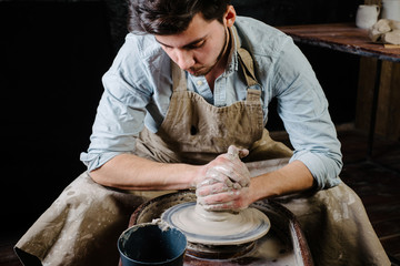 pottery, workshop, ceramics art concept - man working on potter's wheel with raw clay with hands, a male brunette sculpt a utensils near wooden table with tools, master in apron and a shirt
