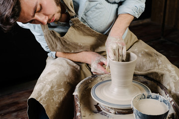 pottery, workshop, ceramics art concept - man working on potter's wheel with raw clay with hands, a male brunette sculpt a utensils near wooden table with tools, master in apron and a shirt