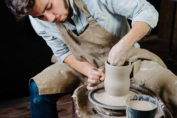 pottery, workshop, ceramics art concept - man working on potter's wheel with raw clay with hands, a male brunette sculpt a utensils near wooden table with tools, master in apron and a shirt