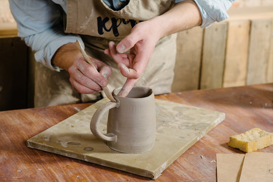 potter, workshop, ceramics art concept - ceramist holding cup and carefully examines the product, young male master dressed in a stained clay apron, craftsman's hands holding unbaked jug