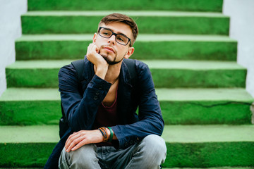 Obraz premium Portrait of a handsome teen student guy in glasses sitting on the green sairs - education, hischool and people concept.