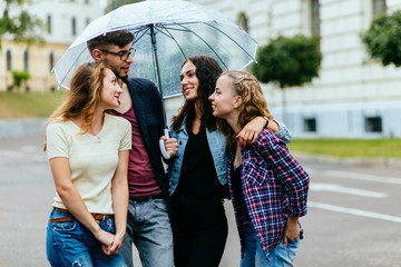 Group of four positive student friends sheltering from rain beneath a big umbrella - education, school, teamwork and people concept.