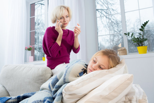 Mother Measuring Temperature Of Her Child And Calling Doctor. Sad Mom Holding Thermometer In One Hand And Cell Phone In Another.