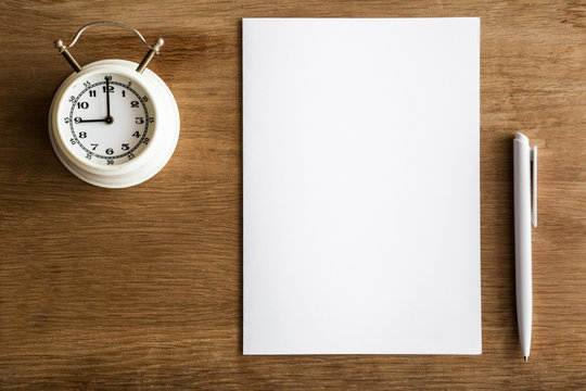 White paper with pen and clock on the wooden table. Examination time. Education concept. Top view. Empty place for a text.