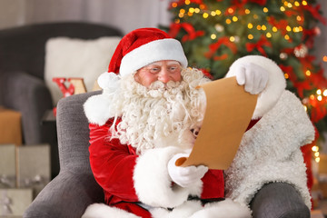 Authentic Santa Claus with sheet of paper sitting in armchair at room decorated for Christmas