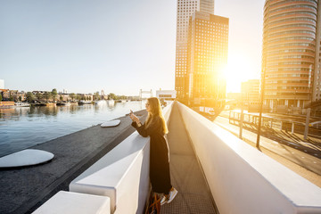 Lifestyle portrait of a stylish woman standing on the modern bridge with skyscrapers on the background during the morning in Amsterdam city