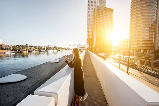 Lifestyle portrait of a stylish woman standing on the modern bridge with skyscrapers on the background during the morning in Amsterdam city