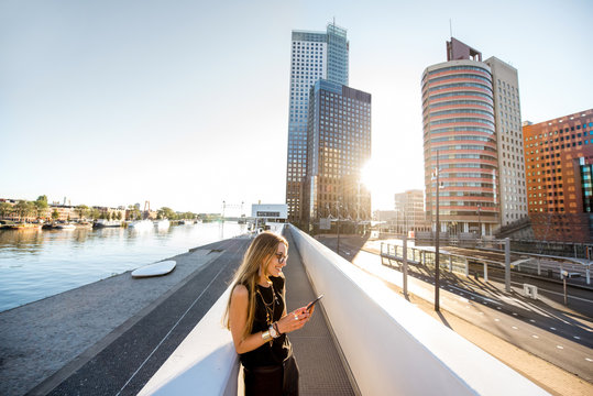 Lifestyle portrait of a stylish woman standing with phone on the modern bridge with skyscrapers on the background during the morning in Amsterdam city